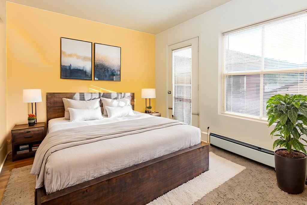 A bedroom with a bed, two lamps, a potted plant, and a painting on the wall.