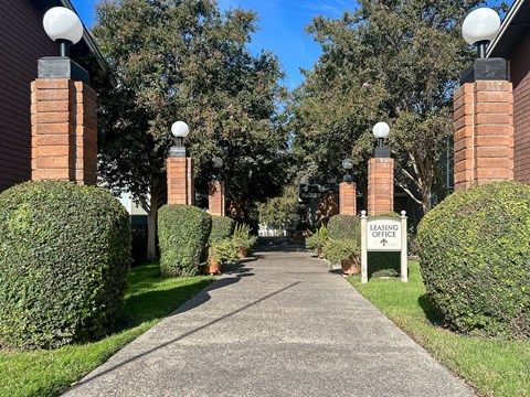 A pathway leads to a leasing office surrounded by greenery and brick pillars with spherical lights.