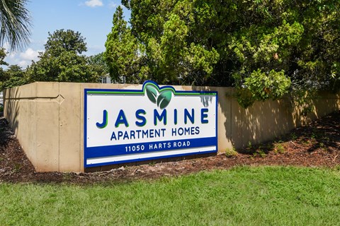 A sign for Jasmine Apartment Homes is displayed in front of a wall and trees.