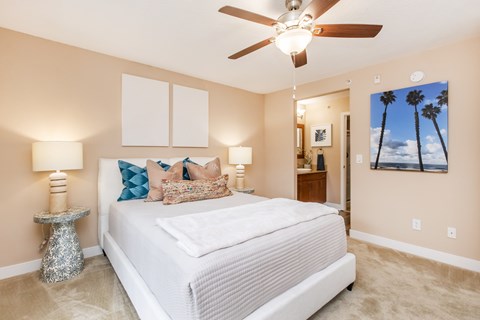 A bedroom with a large bed, a ceiling fan, and a painting of palm trees on the wall.