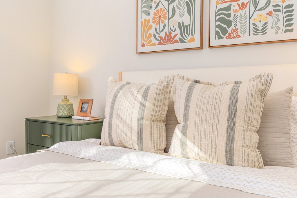 A bed with a white comforter and striped pillows is in front of a wall with two framed artworks.