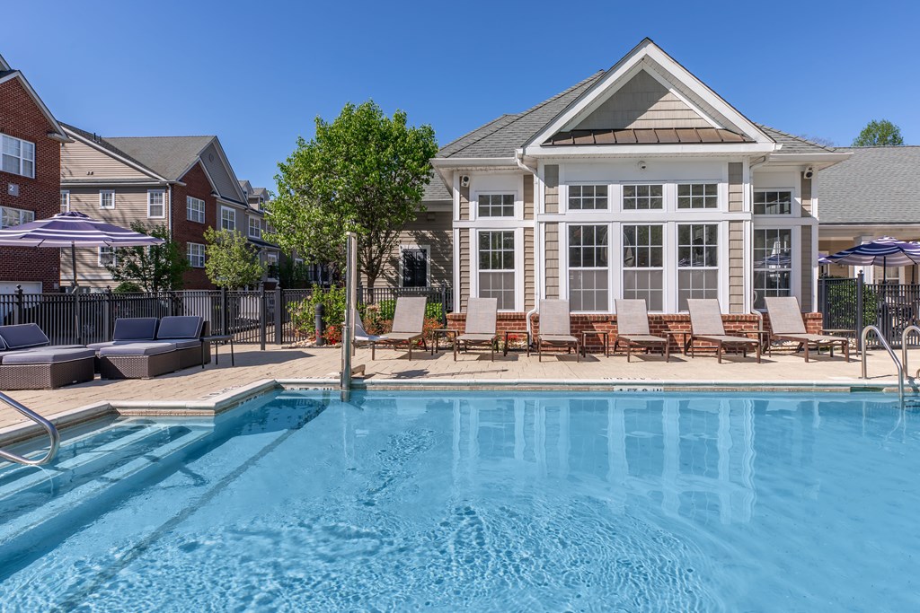 A swimming pool in front of a house with a patio and chairs.