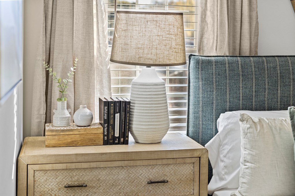 A lamp with a beige shade sits on a wooden nightstand next to a blue striped pillow at West Oak Apartments, North Carolina