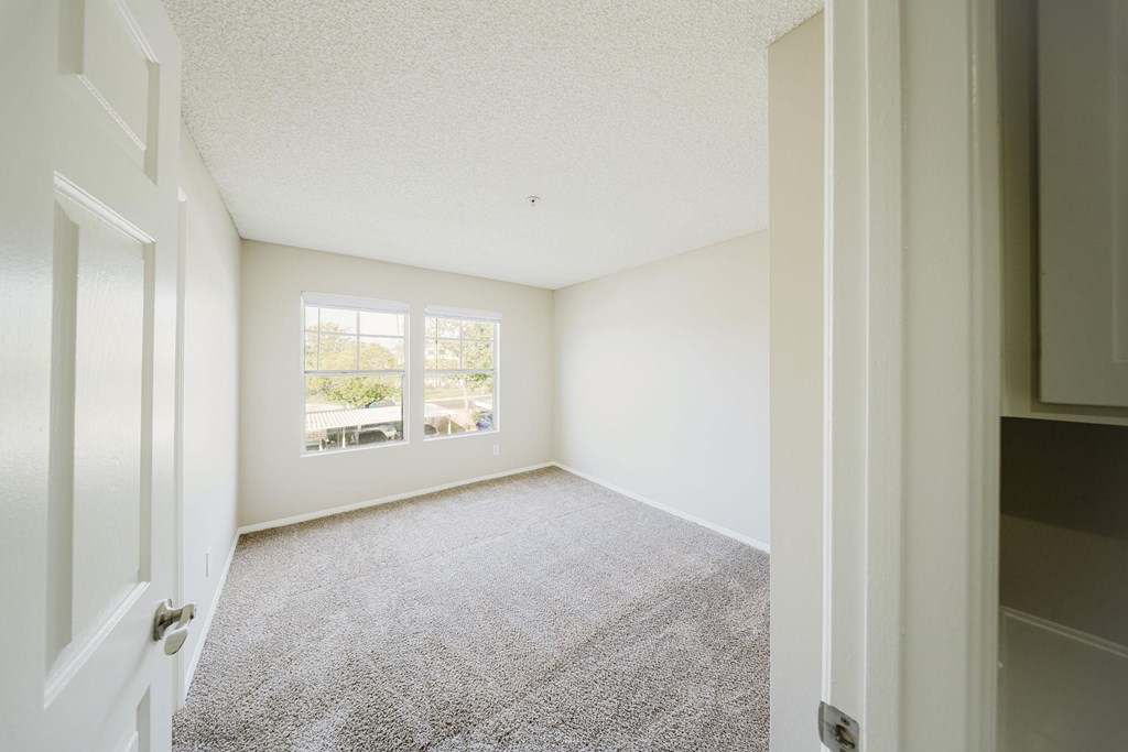 A room with a carpeted floor and a window with a view of trees and a balcony. at Legends at Rancho Belago, Moreno Valley, CA, 92553