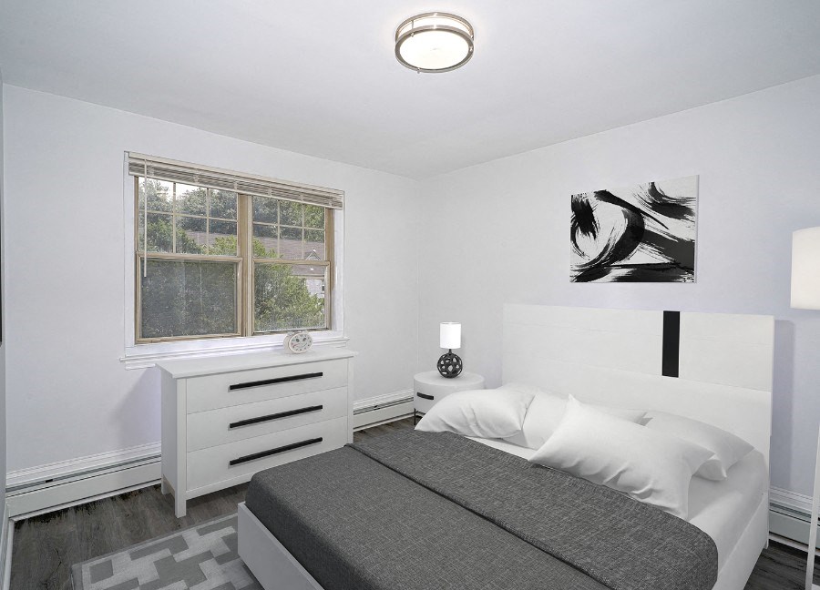 A bedroom with white walls and gray-brown vinyl floors. There is a full-size bed with a white headboard and gray comforter. A black and white painting is on the wall above the bed, and a white dresser is against the wall to the left under a window.