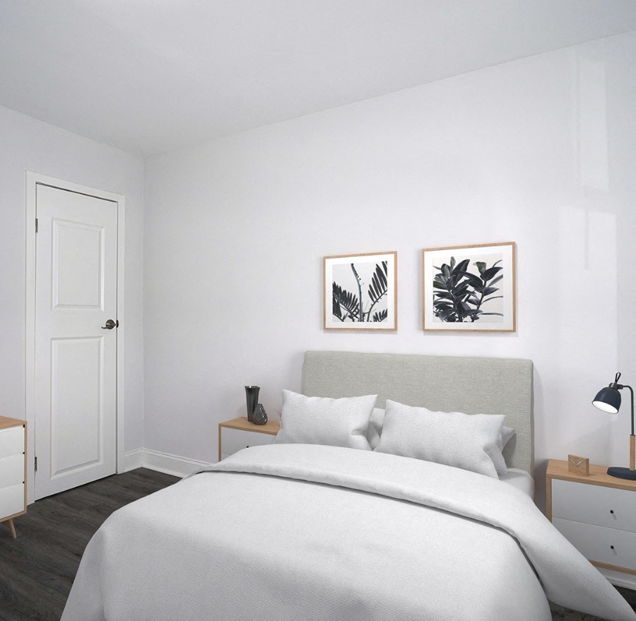 A small bedroom with white walls and gray-brown vinyl floors. The bed has a light gray headboard and a white comforter and pillows. Two foliage prints hang on the wall above the headboard. Small end tables are on either side of the bed, and one has a lamp on it. The door is on the wall to the left of the bed.