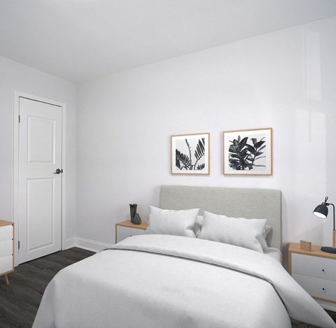 Bedroom at Drumcastle Apartments A small bedroom with white walls and gray-brown vinyl floors. The bed has a light gray headboard and a white comforter and pillows. Two foliage prints hang on the wall above the headboard. Small end tables are on either side of the bed, and one has a lamp on it. The door is on the wall to the left of the bed.