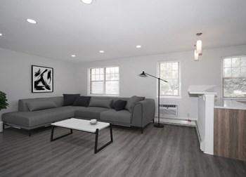 Furnished Living Room at Drumcastle Apartments Living room with white walls and gray-brown vinyl floors. An L-shaped couch is in the left corner of the room, with a black and white painting on the wall above one side of the couch, and two windows behind the other side. The couch is facing a white coffee table. The white kitchen counter is to the right - Photo Gallery 3