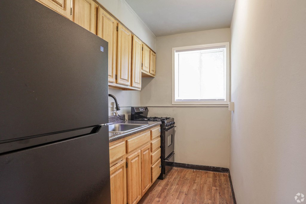 View of the kitchen with a black fridge, dark countertops, light brown cabinets, brown wooden floors, and a small window
