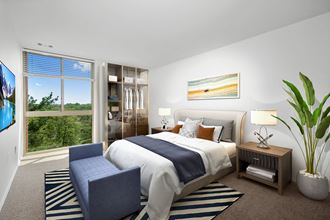 Furnished bedroom with white walls, a big window on the far wall next to a stand-alone closet. There is a white bed on a zebra-patterned area rug. There is a blue accent blanket across the middle of the bed and a blue bench at the foot of the bed. There are brown end tables with lamps on either side of the bed, and a big plant in the corner.