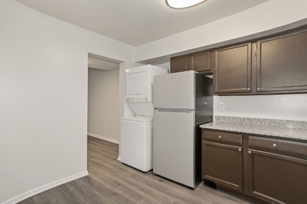 Kitchen with white walls and light brown vinyl floors. The kitchen cabinets are dark brown and the countertop is a light speckled color. The fridge is next to the countertop, and there is a stacked washer and dryer immediately to the left of the fridge.