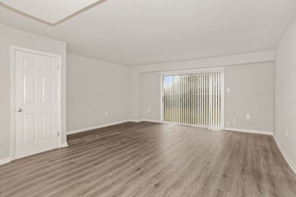 Unfurnished living room with white walls and light brown vinyl floors. There is a white door on the wall to the left, and a glass sliding door on the back wall that leads outside.