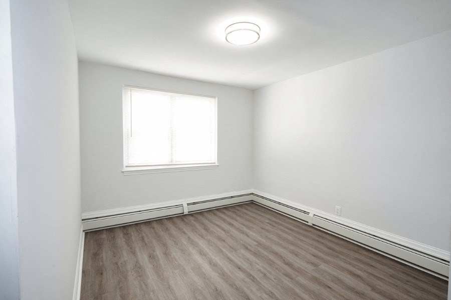 An unfurnished small bedroom with white walls and vinyl flooring. There is a window on the back wall of the room and a round light in the center of the ceiling