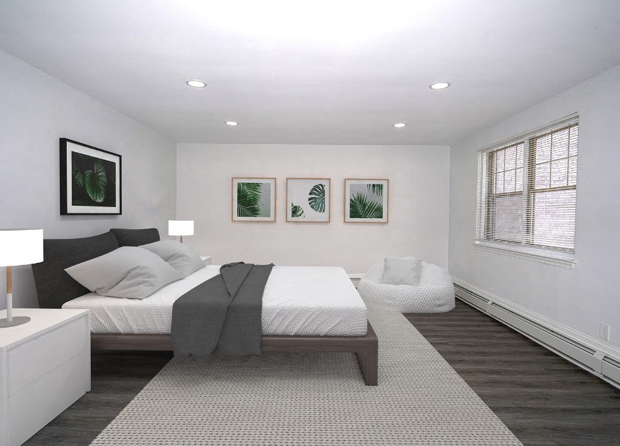 A bedroom with white walls and gray-brown vinyl floors. There is a full-size bed with a white comforter, gray pillows and a gray throw blanket. There are white end tables with lamps on them on both sides of the bed. Across from the bed is a big window