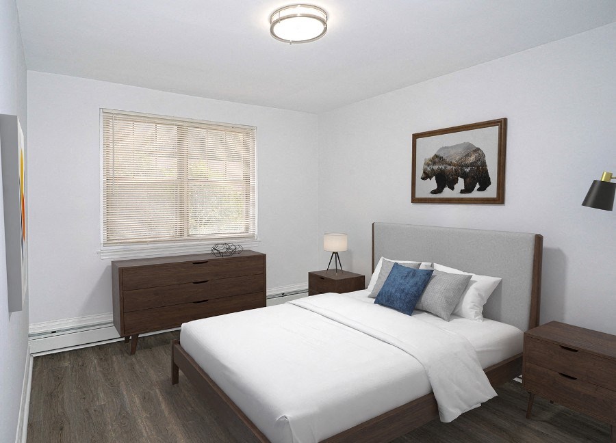 A bedroom with white walls and gray-brown vinyl floors. There is a full-size bed with a gray headboard and white comforter. A bear painting hangs on the wall above the headboard. There are wooden end tables on both sides of the bed and a dresser against the wall to the left under a window