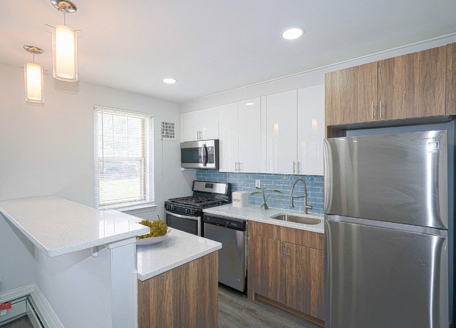 kitchen with blue tile backsplash and stainless-steel appliances. The walls are white and the floor is gray-brown vinyl. Cabinets above the fridge are brown and the cabinets under the countertops are brown.