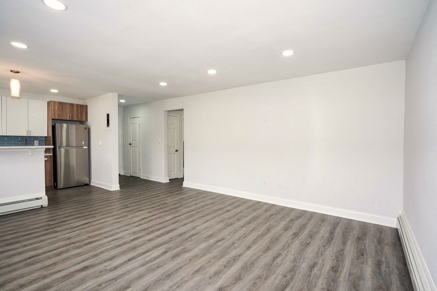 Unfurnished living room with white walls and gray-brown vinyl floors. The stainless-steel fridge and entrance to the kitchen can be seen at the left end of the photo