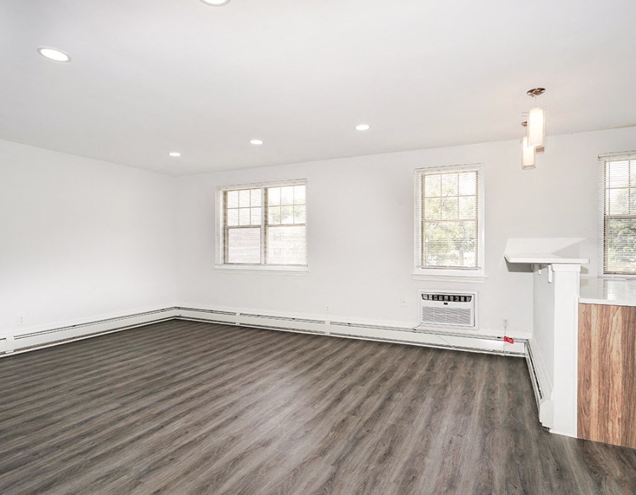Unfurnished living room with white walls and gray-brown vinyl floors. There are three windows on the back wall of the living room, and the kitchen counter is to the right.