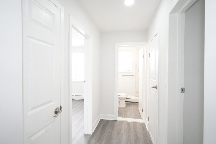 The view of a hallway inside an apartment. There are doors leading to rooms on both sides of the hallway. The walls and doors are white, and the floor is gray-brown vinyl.