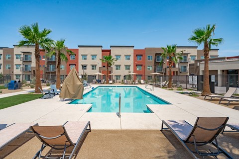 A swimming pool surrounded by palm trees and lounge chairs in front of a multi-story apartment building.