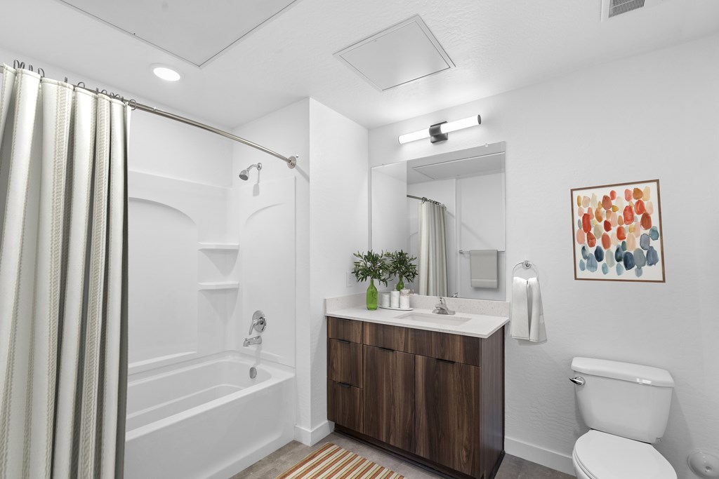 A bathroom with a white toilet, a white sink, a white bathtub, a wooden cabinet, a mirror, a plant, and a painting.