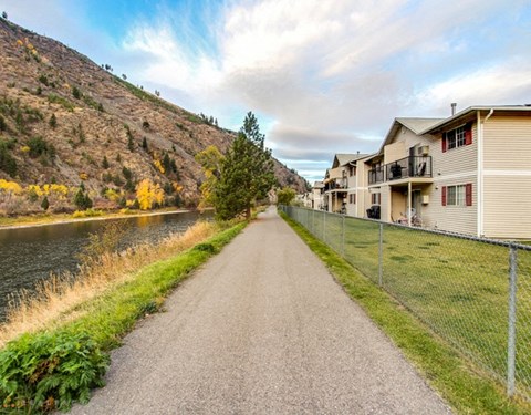A pathway runs alongside a river with houses on one side and a hill with trees on the other.