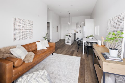 A brown leather couch with white pillows sits in a living room with a white rug and a wooden table.