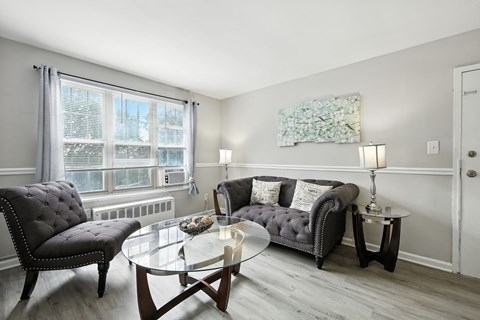A living room with a grey couch, a black chair, and a glass coffee table at Huntwood Courts Apartments, Washington DC