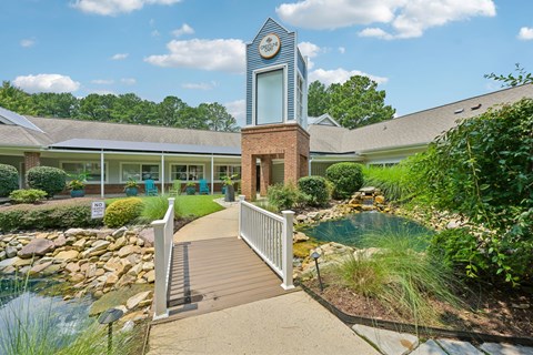 A small building with a clock tower is surrounded by a garden and a bridge.