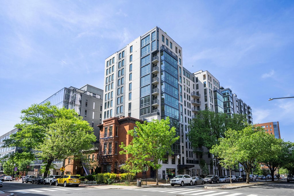 A modern multi-story building with a glass facade is surrounded by trees and other buildings.