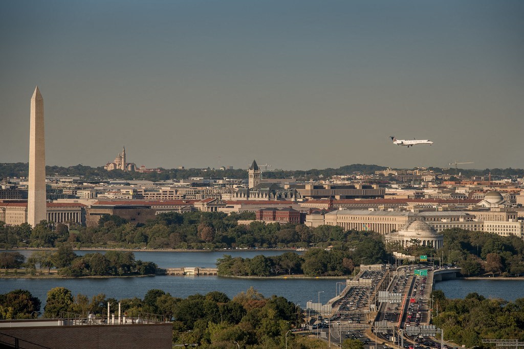 a plane flies over the capital of the united states with the washington monument in the foreground at The Acadia at Metropolitan Park, Arlington, 22202