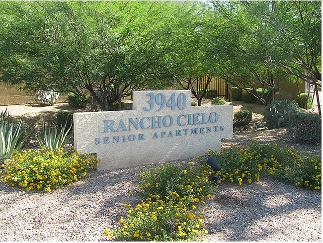 A sign for Rancho Cielo Senior Apartments is surrounded by flowers and greenery at Rancho Cielo Senior Apartments, Phoenix, Arizona
