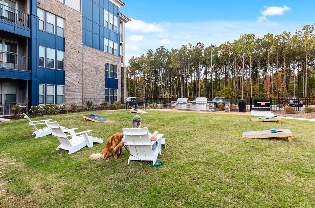 A person is sitting on a white chair in a grassy area with a dog and a picnic table.