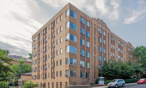 A tall brick building with many windows and cars parked in front.