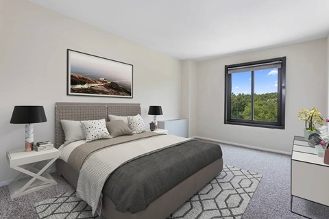 A bedroom with a large bed, a nightstand with a lamp, a framed picture on the wall, and a window with a view of trees.