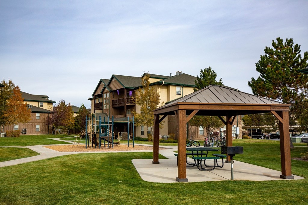 A wooden pavilion with a picnic table sits in a grassy area in front of apartment buildings.at Sunset Ridge, West Jordan