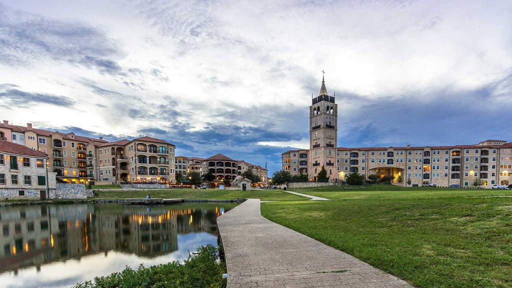Looking along a sidewalk at dusk - to the left is the lake, to the right is an open field. Ahead the Bell Tower and various stone building of Adriatica can be seen.
