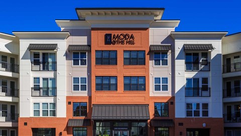 The front entrance to the apartment building. There is red brick in the middle with the Moda at The Hill logo sign at the top. The building is white and grey to the left and right.
