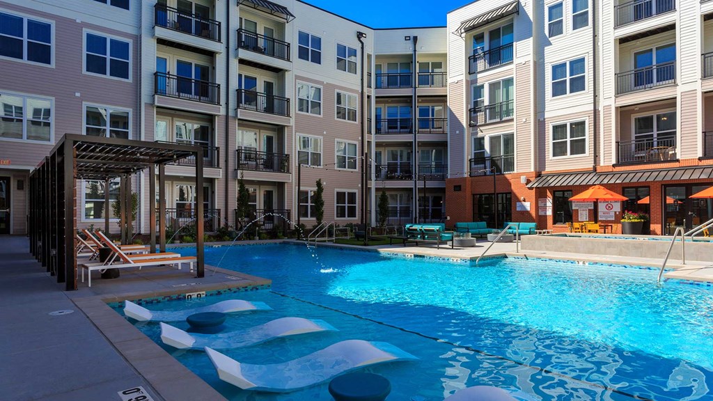 Standing at the edge of the outdoor pool. In-water lounge chairs are just ahead with cabanas just beyond. The apartment building stands tall in the background.