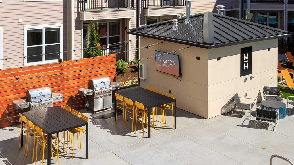 There are two grills against a wooden wall with high top tables in front. A small building is on the right with an outdoor television mounted on it.