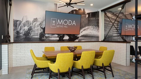 A lounge seating area with a dining table surrounded by yellow chairs. There is a large television mounted on the wall behind.