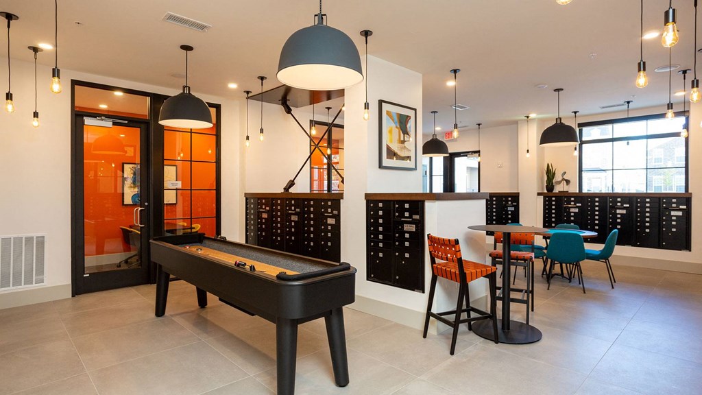 The mail room doubles as a game room. A shuffleboard table is in the foreground with gaming tables off the right. There are mailboxes built-in throughout the room.