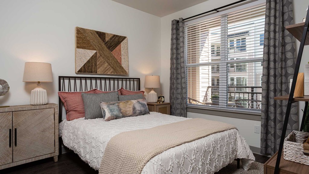 A residence bedroom with a large bed set nicely in the middle, a night stand on both sides. The balcony is seen outside the window.