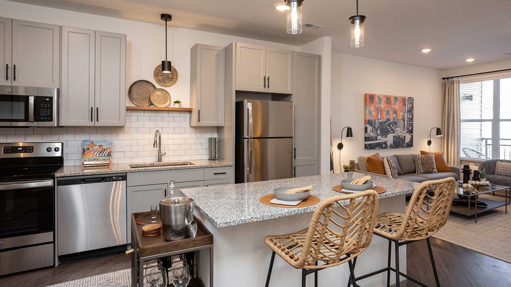 The kitchen is on the left with grey cabinets and stainless steel appliances. The living room is off to the right with a couch against the wall beneath a large art piece.