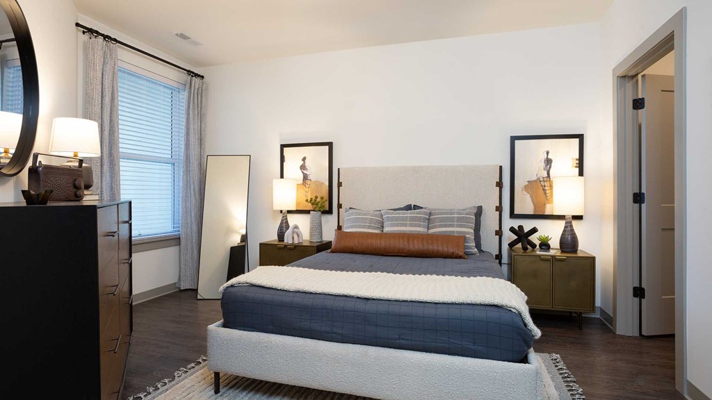 Standing at the end of a large bed with end tables on both sides. There is a dresser and window on the left and the door to another room on the right.