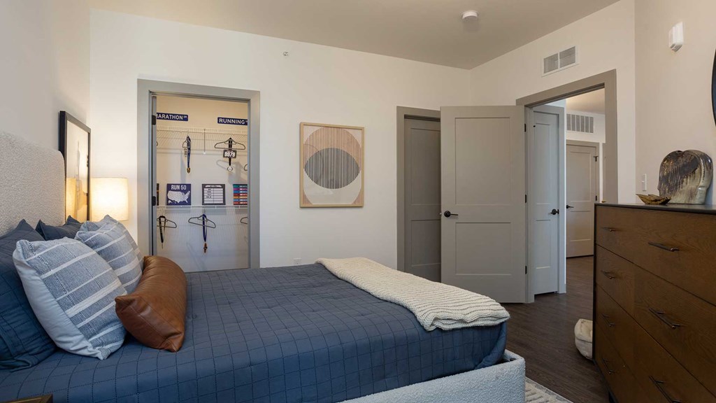 Standing next to a large bed. There is a closet door open across the bed on the left and an open door to the rest of the residence on the right.