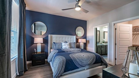 A modern bedroom with a navy blue accent wall, white tufted headboard, and matching blue bedding. two round mirrors hang above bedside tables, and an open door leads to a bathroom.