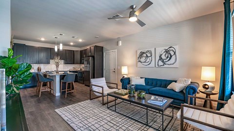 Modern apartment living room with a blue sofa, white chairs, dark kitchen cabinets, and contemporary decor.