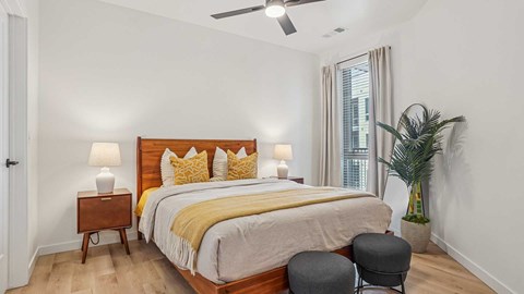 Modern apartment bedroom with wood bed frame, neutral bedding, bedside lamps, wood-style flooring, and large window with natural light.