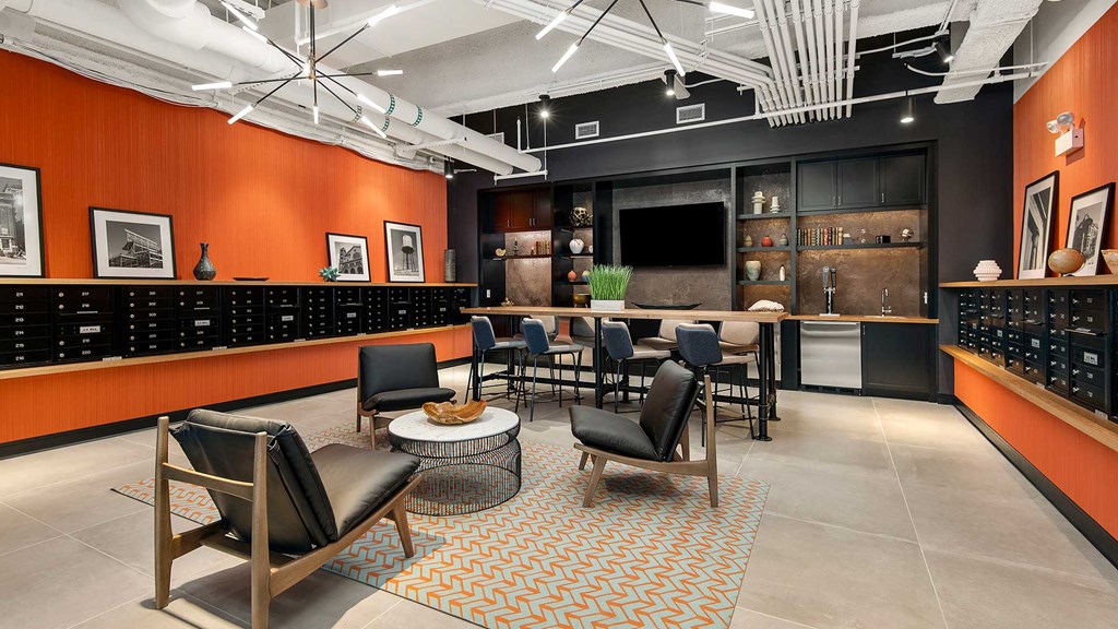 Looking in to the Clubroom at Wrigleyville Lofts. There are club chairs in a circle ahead with a high-top table behind. The far wall has a mounted television with beer taps beside. The side walls are orange with mailboxes built-in for residents.